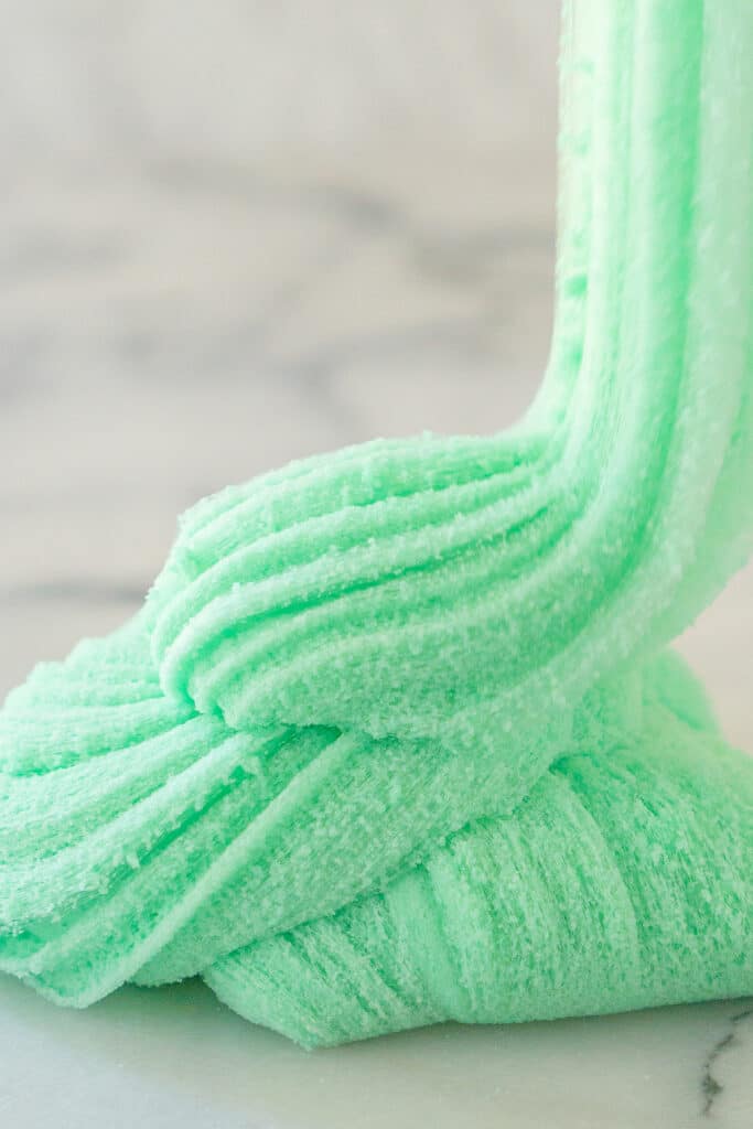 An incredibly fluffy ribbon of sea foam green cloud slime drizzles down into a swirly pile on a white marble counter. The texture of the slime looks like spun cotton candy.