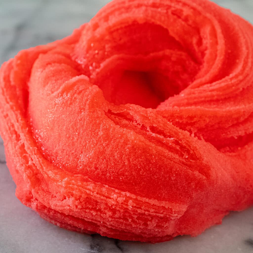 A swirl of bright red icee slime on a white and grey marble counter. The slime texture is wet with fluffy snow particles just visible.
