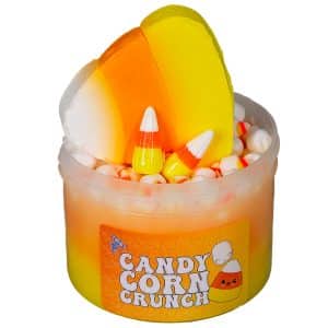 A clear jar is layered with yellow, orange, and white slime behind a label that says Candy Corn Crunch Slime. The slime is topped with a large clay candy corn, two realistic resin candy corn charms, and a generous amount of fimo candy corn sprinkles. Styrofoam mini marshmallows are visible below the toppings, mixed into the white layer of slime.