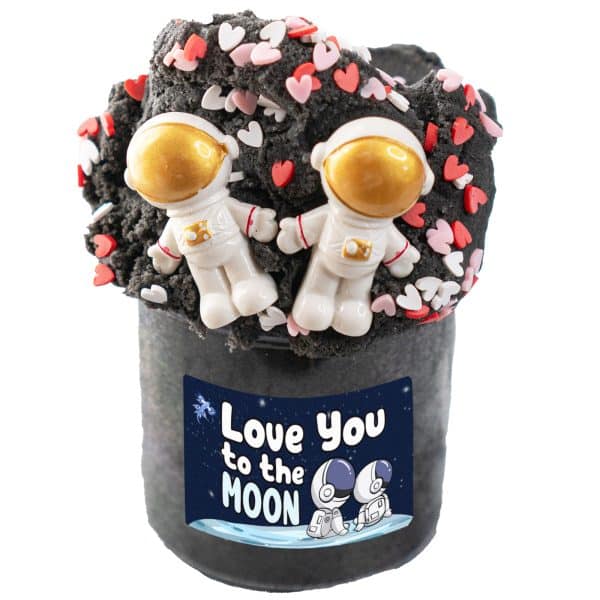 A clear jar is filled high above the top with a dark gray, almost black, fluffy space slime. The top of the cloud dough slime is covered in red, pink, and white heart sprinkles, with two astronaut charms nestled together in front. The label reads, love you to the moon.