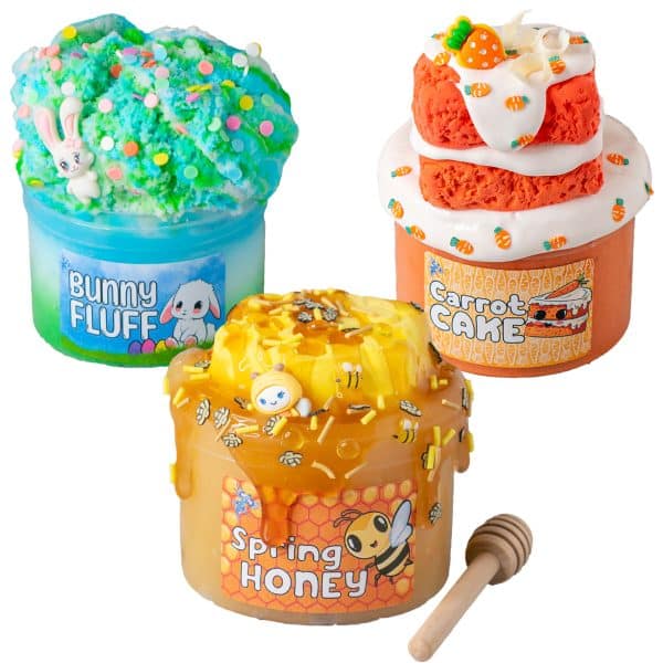 3 Easter slimes on a white marble board. In front is a golden slime that looks like a jar of honey topped with a clay honeycomb. The back right is a fluffy green, white, and blue cloud slime with a cute bunny on it. The back right is an orange cloud slime with white glossy slime that looks like frosting dripping down from orange carrot cake clays.