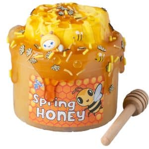 A jar is filled with a pale golden jelly slime that looks just like raw honey. On top is a yellow claw honeycomb drizzled with a brown clear slime that looks like honey dripping down. fake bees and yellow sprinkles are scattered across the top. A wooden honey dipper sits nearby and a label on the jar reads Spring Honey Slime.