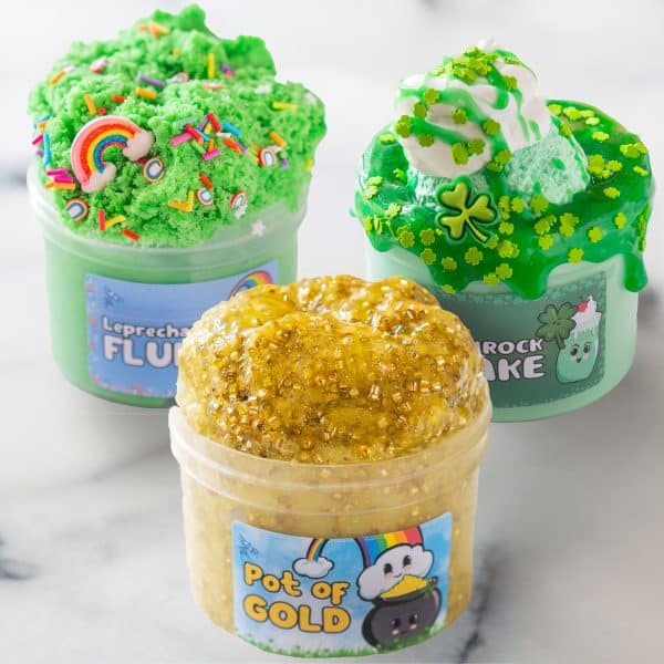 Three translucent plastic jars are filled and piled high with three different St. Patrick's Day slimes. The front jar has a gold clear slime packed with gold beads and glitter. The back left jar has a fluffy green cloud slime covered in rainbow sprinkles and charms. The back right jar is a DIY clay kit with a minty green cloud dough slime topped with green clear slime that looks like syrup, a clay that looks like an ice cream scoop, a white butter slime that looks like whipped cream, and is covered in shamrock sprinkles and charms.