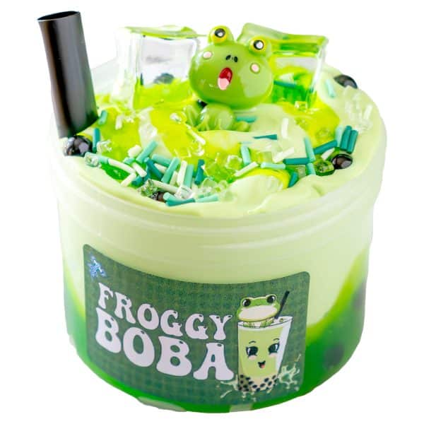 A clear plastic container labeled "Froggy Boba Slime," is filled with swirled light and dark green slime designed to resemble a cup of boba tea. The top of the slime is decorated with a cheerful green frog figurine, clear faux ice cubes, sprinkles, and black beads that mimic boba pearls. A black straw is also inserted into the slime, completing the boba tea illusion.