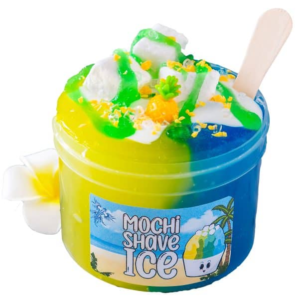 A vibrant yellow and blue Icee Slime looks just like a tropical shave ice, and a label on the jar reads "Mochi Shave Ice Slime". The split colored slime is topped with realistic mini mochi clay pieces, drizzles of green slime that looks like syrup, and white slime that looks like sweetened condensed milk. A pineapple charm, small wooden spoon and a delicate plumeria flower accentuate the playful, summery theme.