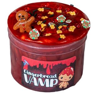 A jar of deep red, shimmery clear slime resembles blood and is topped with a cute vampire gingerbread man charm. The surface is also decorated with gingerbread-themed sprinkles, featuring mini gingerbread houses and gingerbread men, alongside darker vampire-themed accents for a spooky holiday twist. The label on the jar reads Gingerbread Vamp Slime.