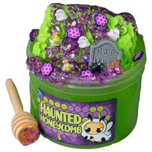 A jar of bright green slime labeled "Haunted Honeycomb" features a neon green honeycomb-shaped clay piece on top, coated in a glittery purple slime that resembles dripping honey. The decorations include ghost and bee charms, a tombstone, and Halloween-themed sprinkles, evoking the eerie concept of bees making honey in a spooky cemetery.