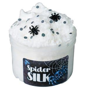 A fluffy white cloud slime is packed into a jar, while the stretched out slime with a soft, airy texture is drizzled perfectly on top to resemble delicate spider webs. Black spider charms and tiny spiderweb sprinkles decorate the surface, adding a spooky touch, while the label reads "Spider Silk" with a cute blue spider and spiderweb.