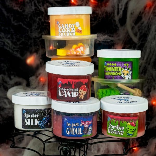 6 Halloween cute and spooky slimes are displayed on a black cake stand that looks like a spiderweb with a smokey black and orange background.